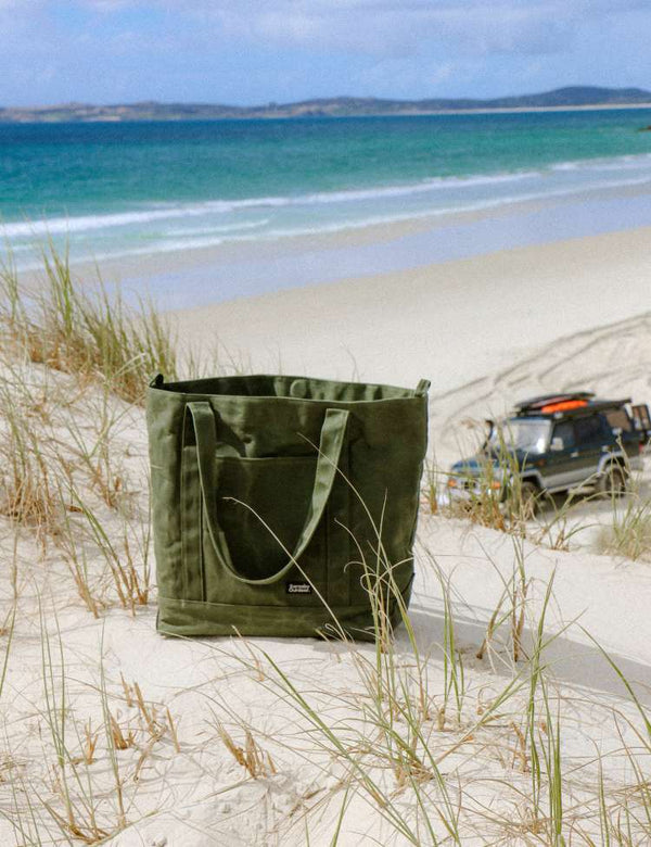 Green Barnaby Better tote bag on a sandy beach with ocean and vehicle in the background
