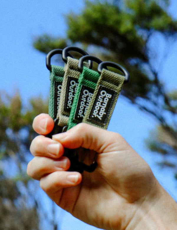 Hand holding four green Trail Key Clip keychains with brand name against a blurred natural background