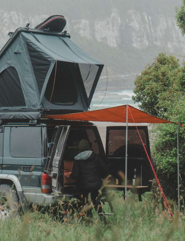 Van with rooftop tent and open door in a natural setting utilising a waxed canvas mat as rain cover.