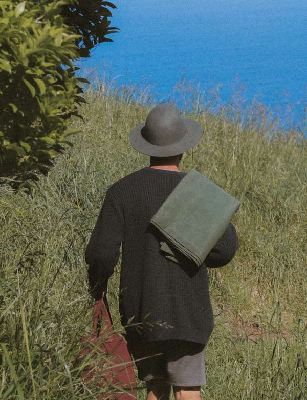 Person walking on a grassy hill with a blue sky and ocean in the background carrying a waxed canvas Barnaby Mat