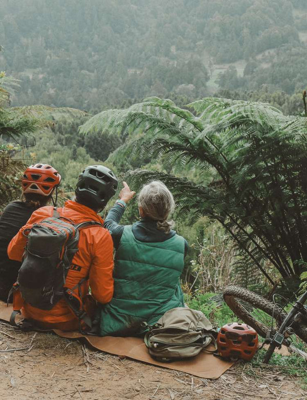 Three mountain bikers are sitting on a waxed canvas mat in a forest, pointing at something in the distance.