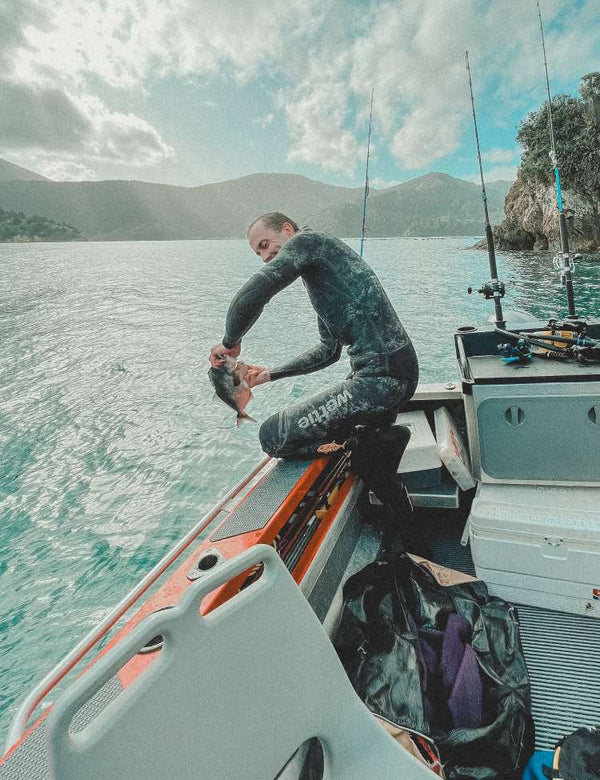 Person in a spearfishing wetsuit on a boat holding a fish with mountains and water in the background
