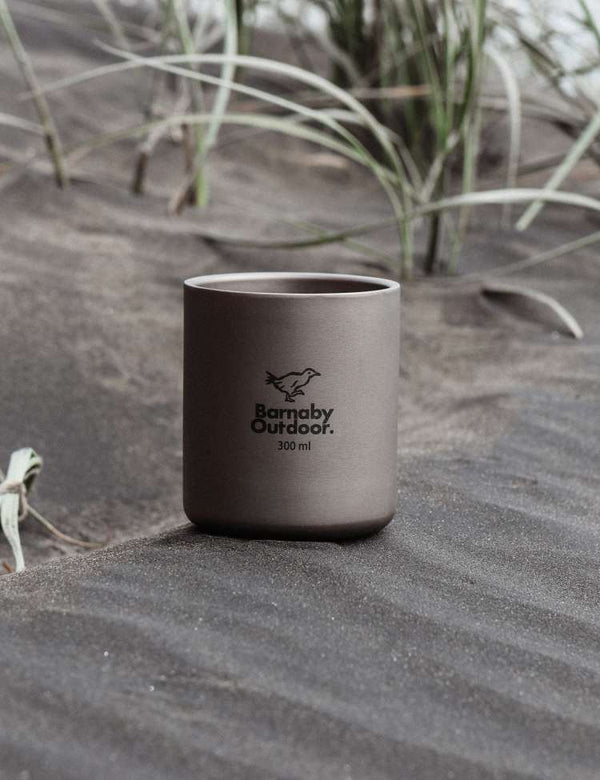 Dark gray titanium mug with 'Barnaby Outdoor' logo on sand