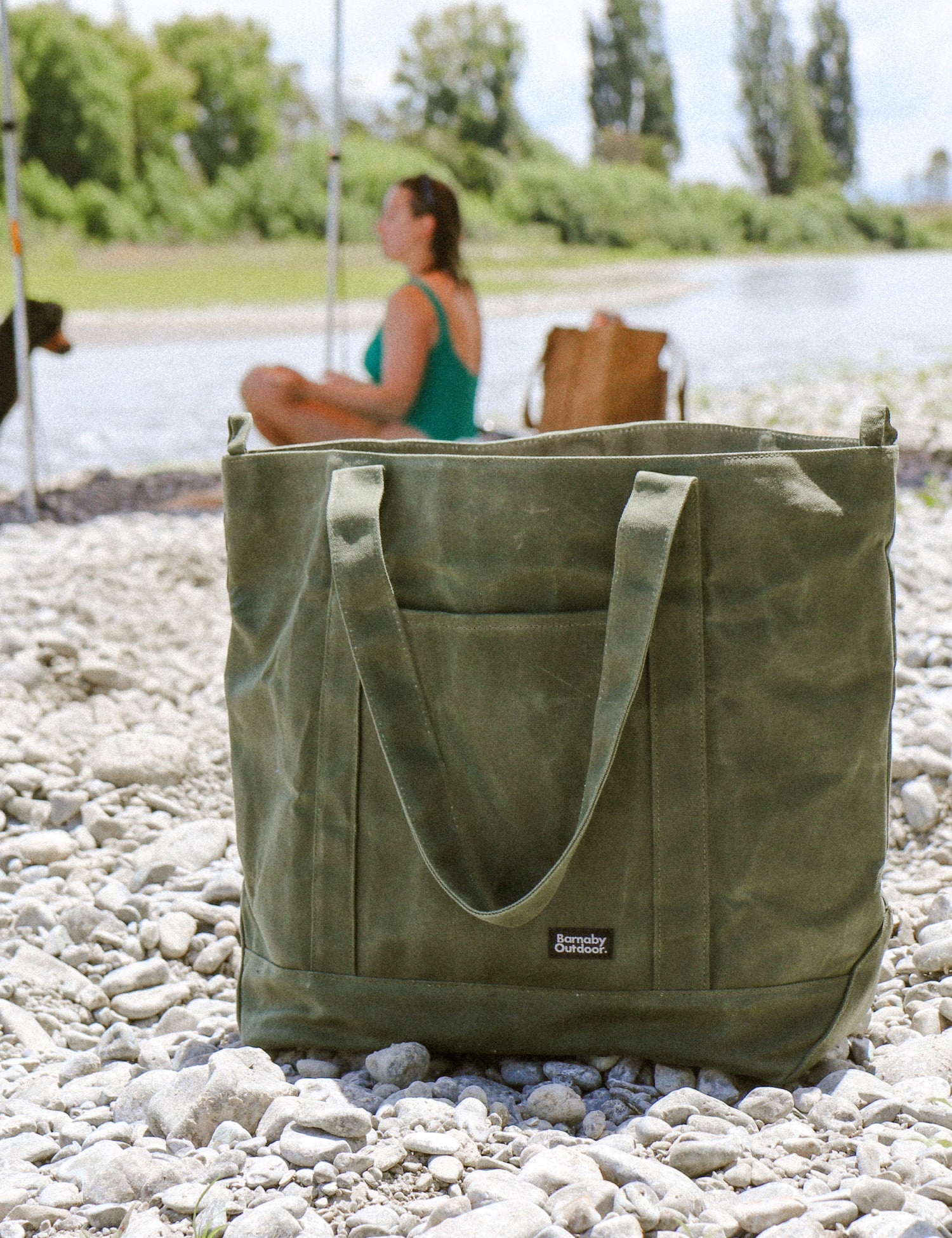 Green tote bag on pebbles with a person sitting by a lake in the background