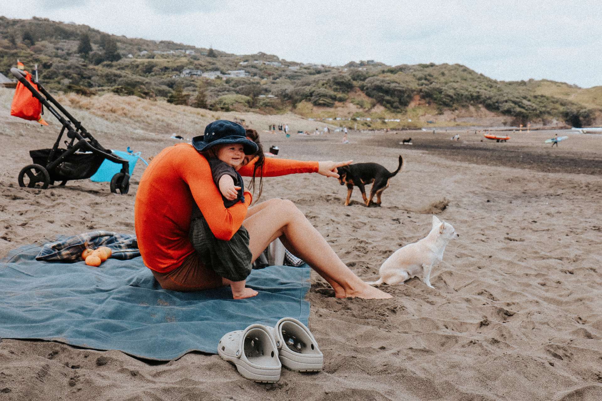 Mother and baby sitting on a beach with two dogs on a waxed canvas mat, surrounded by a stroller and beach gear.