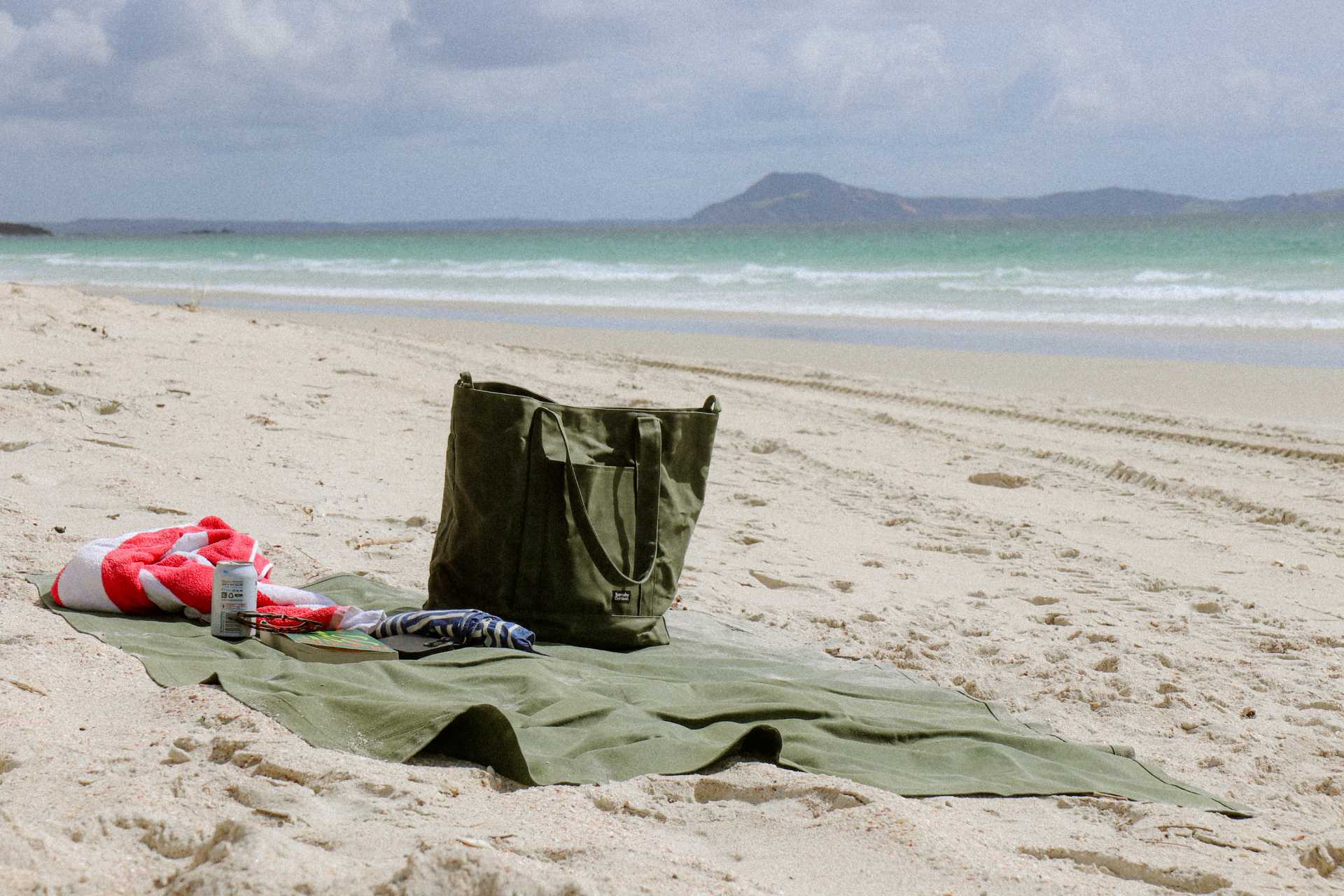 Green waxed canvas mat on a sandy beach with a waxed canvas tote bag and water bottle nearby, under a cloudy sky.